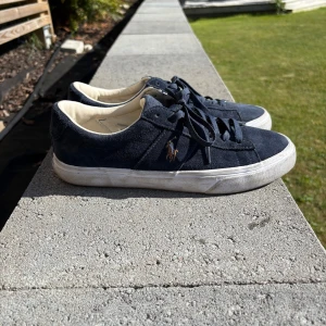 Mörkblå sneakers från Polo Ralph Lauren - Säljer ett par mörkblå sneakers från Polo Ralph Lauren med klassisk design. Skorna har snörning, vit platt sula och är tillverkade i mocka. Polo-logga på sidan och bak på sulan. Perfekta för en stilren vardagslook eller för finare tillfällen 