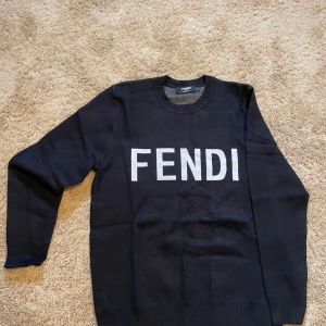 Svart stickad tröja från Fendi - Säljer en svart stickad tröja från Fendi med stor vit logotyp på bröstet. Tröjan har rund halsringning, långa ärmar och ribbade muddar vid ärmslut och nederkant. Perfekt för dig som gillar stilrena märkesplagg. står M i nacken men passar som en svensk klassisk S