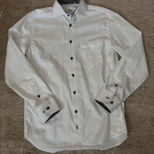 Vit skjorta med mönstrad krage från Oscar of Sweden - Snygg vit skjorta från Oscar of Sweden med svartvit blommig insida på krage och manschetter. Klassisk passform, långärmad och med svarta knappar. Perfekt för dig som vill ha en stilren look med en twist.