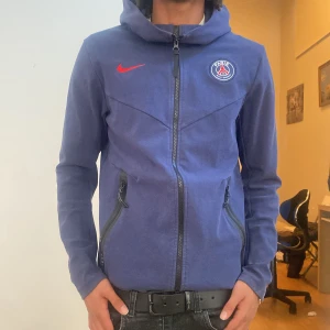 Blå PSG hoodie från Nike - Säljer en blå hoodie från Nike med Paris Saint-Germain-logga på bröstet och röd Nike-swoosh. Jackan har dragkedja, huva och två fickor med dragkedja. Perfekt för fotbollsfans som vill ha en snygg och sportig look.