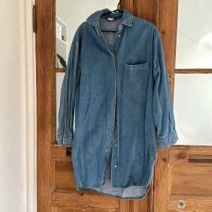 Jeansklänning från &Denim - Säljer en klassisk jeansklänning från &Denim i ljusblå denim. Klänningen har lång ärm, bröstficka och knäppning framtill. Perfekt för en avslappnad och cool stil.