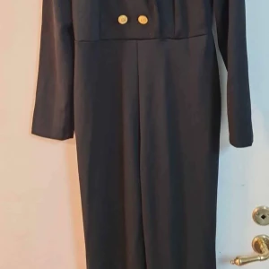 Svart långklänning med guldknappar - Säljer en svart byxlinning med långa ärmar och snygg krage. Byxorna har dekorativa guldknappar framtill och en klassisk, stilren look. Perfekt för dig som gillar en elegant och tidlös stil.