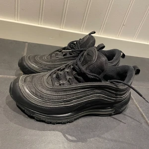 Svarta Nike Air Max 97 sneakers - Säljer ett par svarta Nike Air Max 97 sneakers med vågformade detaljer och synlig luftbubbla i sulan. Skorna har snörning och är tillverkade i syntetmaterial och textil. Perfekta för dig som gillar en sportig och stilren look.