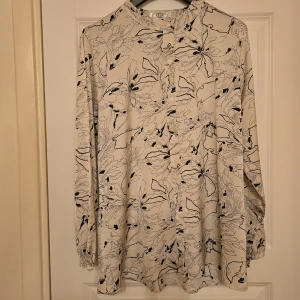 Vit blus med svart blommigt mönster - Säljer en vit blus med svart blommigt mönster. Blusen har långa ärmar med resår vid ärmslut och knappar framtill. Tillverkad i mjuk viskosblandning med lite stretch för extra komfort. Perfekt för dig som gillar stilrena men ändå unika plagg.