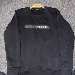 Svart sweatshirt från Philipp Plein - Säljer en svart sweatshirt från Philipp Plein med logotyp i relief på bröstet. Tröjan har rund halsringning och är tillverkad i 100% bomull, vilket gör den mjuk och bekväm. Perfekt för en stilren och enkel look.