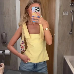 Gul linnetopp med rosettband - Snygg ljusgul linnetopp med breda axelband som knyts i rosetter. Toppen har fyrkantig ringning och figurnära passform. Perfekt till jeans eller kjol till sommaren 💕