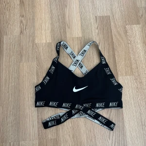 Svart sport-bh från Nike med logoband - Säljer en svart sport-bh från Nike med elastiska band med Nike-logga i vitt. Korsade axelband och bred resår under bysten för extra stöd. Perfekt för träning och sportiga aktiviteter. Storlek S. Pris 50kr