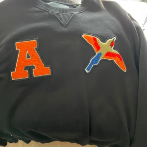 Svart sweatshirt med applikationer från Axel Arigato - Säljer en svart sweatshirt från Axel Arigato med en stor orange bokstav 'A' och en färgglad fågelapplikation på bröstet. Tröjan har rund halsringning och ribbade muddar. Perfekt för dig som gillar streetwear och unika detaljer. 