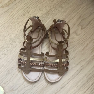 Bruna sandaler med flätade detaljer från H&M - Säljer ett par bruna sandaler från H&M med flera remmar i olika nyanser av brunt och guld. Skorna har både släta och flätade remmar samt dragkedja bak för enkel påtagning. Perfekta för varma dagar. Skorna ger dig en snygg och cool look!