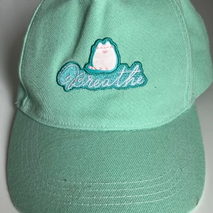 Mintgrön keps med broderad pusheen katt - Snygg mintgrön keps från CultureFly med broderad katt och texten 'Breathe' framtill. Justerbar rem baktill för passform. Tillverkad i 100% polyester