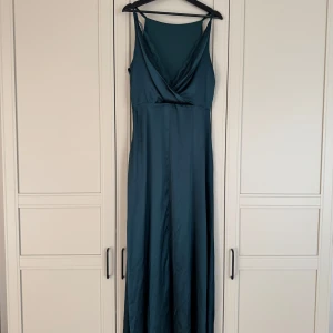 Mörkgrön långklänning från Bubbleroom Occasion - Säljer en elegant mörkgrön långklänning från Bubbleroom Occasion. Klänningen har omlottliv med spetsdetaljer på ryggen, smala axelband, slit snyggt vid benen och är tillverkad i ett glansigt satinliknande material. Perfekt för speciella tillfällen som bal eller finmiddag! Originalpris är 1200kr, riktigt kap!