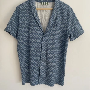 Blå kortärmad skjorta från Morris - Säljer en blå kortärmad skjorta från Morris med vitt geometriskt mönster. Skjortan har klassisk krage och knappar framtill. Perfekt för varma dagar och enkel att matcha med jeans eller shorts.