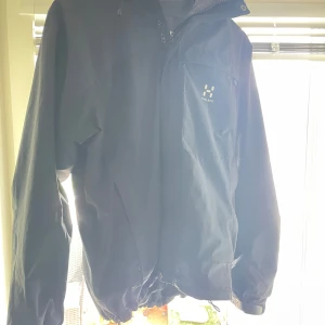 Svart vindjacka från Haglöfs - Säljer en svart vindjacka från Haglöfs med huva och dragkedja. Jackan har en bröstficka med logga och justerbar nederkant. Perfekt för blåsiga dagar och enkel att packa med sig.