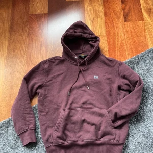 Vinröd hoodie från Lee - Säljer en vinröd hoodie från Lee med klassisk huva och dragsko. Tröjan har en stor magficka framtill och diskret Lee-logga på bröstet. Perfekt för en avslappnad stil och enkel att matcha med jeans.