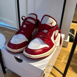 Nike Air Jordan 1 Low röd och vit - Säljer ett par Nike Air Jordan 1 Low i rött och vitt med svarta detaljer. Skorna har klassisk snörning, rund tå och platt sula. Perfekta för dig som gillar streetwear och vill sticka ut med en ikonisk sneaker.