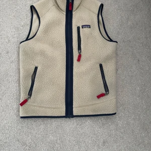Patagonia väst beige - Säljer en beige fleeceväst från Patagonia med svarta detaljer och röda dragkedjor. Västen har tre fickor med dragkedja och hög krage. Perfekt för lager-på-lager och håller dig varm under kyliga dagar. Perfekt till sommarn! 