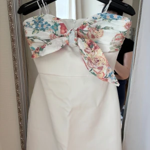 Vit kortklänning med blommig rosett - Supersöt vit kortklänning med axelband och stor blommig rosett framtill. Överdelen har ett blommigt mönster i pastellfärger och klänningen har en feminin, figurnära passform. Perfekt för sommarens alla tillfällen!