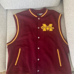 Vinröd collegeväst från H&M x Sex Education - Vinröd väst i college-stil från H&M:s samarbete med Sex Education. Väst med gula detaljer, knappar framtill och broderad M-logga med texten 'Dolphins' på bröstet. Perfekt för en sportig look.
