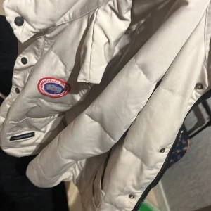 Beige dunjacka från Canada Goose - Säljer en beige dunjacka från Canada Goose med klassisk patch på bröstet. Jackan har långa ärmar, knappar och dragkedja framtill. Perfekt för kyliga dagar och riktigt snygg till streetwear-stilen.