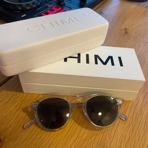 CHIMI glasögon - Snygga solglasögon från CHIMI med transparent båge och runda, mörka glas. Perfekta för att lyfta din stil under soliga dagar. Kommer med originaletui och ask.