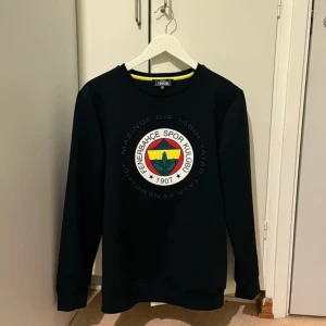 Mörkblå sweatshirt med Fenerbahce-logga - Säljer en mörkblå sweatshirt med Fenerbahce Spor Kulübü-logga tryckt på bröstet. Tröjan har rund halsringning och långa ärmar. Perfekt för dig som vill visa ditt stöd för laget!