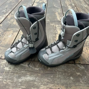 Grå snowboardboots från Salomon - Säljer ett par grå snowboardboots från Salomon. Boots med högt skaft, snörning och robust yttersula. Insidan är vadderad för extra komfort och passformen är anpassad för anatomi. Perfekta för vinterns äventyr i backen.