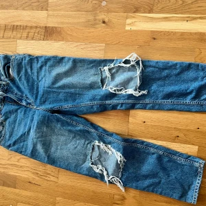 Blå slitna jeans från River Island - Säljer ett par blå jeans från River Island med stora slitningar på båda benen. Jeansen har klassisk femficksdesign och raka ben. Perfekta för en avslappnad look.