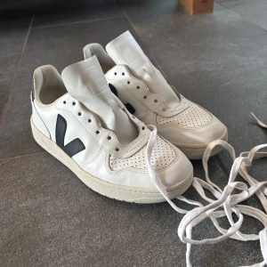 Veja skor - Säljer ett par klassiska vita sneakers från Veja med svart V-logga på sidan. Skorna har perforerad tå, platt sula och snörning. Perfekta till vardags för en clean och stilren look. Endast använda ett fåtal gånger!