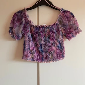 Blommig croppad topp från Amisu - Superfin blommig croppad topp från Amisu i lila, rosa och blå toner. Toppen har puffiga korta ärmar och smockad resår över hela kroppen. Perfekt för varma dagar och ger en söt och trendig look.
