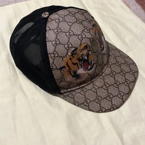 Gucci tigger - Säljer en snygg keps från Gucci med klassiskt GG-mönster och broderade tigrar framtill. Kepsen har svart mesh baktill och justerbar rem med röd-gröna detaljer. Sällsynt modell ligger på runt 5000pluss ny 8 av 10 skick och den är äkta. Dom vita fläckarna på insidan är tvättmedel och inget smuts går att få bort. (Grattis byxor tröja och eller t shirt på köpet om man vill)
