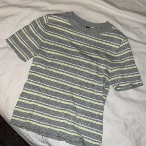 Grå ribbad randig t-shirt - Säljer en grå ribbad t-shirt med smala vita och ljusgula ränder. T-shirten har rund halsringning och korta ärmar. Storlek S