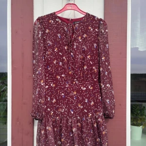 Vinröd blommig klänning från Trendyol - Vinröd klänning med blommigt mönster och långa ärmar från Trendyol. Klänningen har knytning vid halsen, lätt volang nertill och dragkedja i ryggen. Perfekt för dig som gillar romantisk stil och fina detaljer.