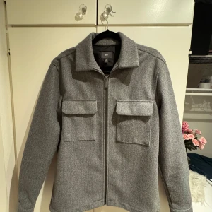 Grå overshirt från H&M - Säljer en stilren grå overshirt från H&M med dragkedja framtill och två stora bröstfickor med lock. Jackan har klassisk krage och raka ärmar, perfekt för lager-på-lager. Enkel och snygg design som passar till mycket. Storlek S
