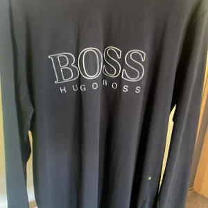Svart långärmad tröja från Hugo Boss - Säljer en svart långärmad tröja från Hugo Boss med stort BOSS-tryck på bröstet. Tröjan är i regular fit och tillverkad i mjuk bomull, perfekt för en stilren och enkel look.