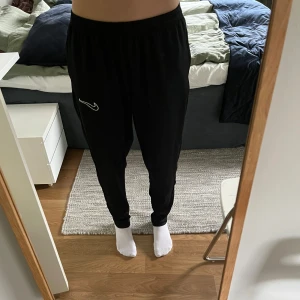 Svarta mjukisbyxor från Nike - Svarta mjukisbyxor från Nike med vit logga på vänster lår. Byxorna har resår i midjan och är raka i modellen med avslappnad passform. Perfekta för chill eller träning.