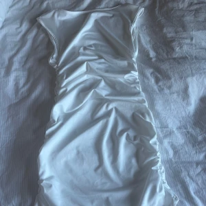 Vit tight kortklänning med rynkade sidor - Säljer en vit tight kortklänning med rynkade sidor. Klänningen är ärmlös och har en enkel, stilren design som passar perfekt till sommaren. Materialet känns mjukt och stretchigt för en bekväm passform. Har en öppen rygg