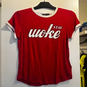 Röd t-shirt med vit text från FB Sister - Röd t-shirt från FB Sister med vit text 'Stay Woke' på bröstet. T-shirten har vita kontrastkanter vid halsringning och ärmslut. Tillverkad i mjuk viskosblandning och har en klassisk passform med korta ärmar.