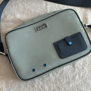 Grå axelväska från Louis Vuitton - Snygg grå axelväska från Louis Vuitton med monogrammönster. Väskan har en justerbar axelrem och en dragkedja upptill. Insidan är klädd i en kontrasterande orange färg. Perfekt för att bära dina dagliga nödvändigheter med stil.