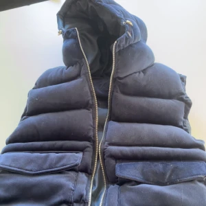 Mörkblå mockaväst med huva - Snygg svart mocka manchester väst med dragkedja och praktiska fickor. Perfekt för kyligare dagar med sin vadderade design och huva för extra värme. Använd 1 gång