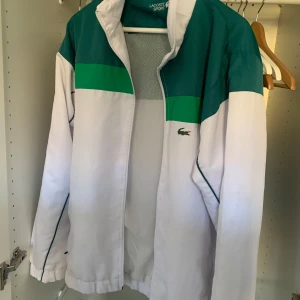 Vit och grön vindjacka från Lacoste - Säljer en snygg vit och grön vindjacka från Lacoste. Jackan har en dragkedja framtill och är långärmad med elastiska muddar. Perfekt för vår och höst med sin sportiga look och klassiska krokodillogga på bröstet.