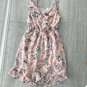 Blommig rosa klänning med volangdetaljer - Söt rosa klänning med blommigt mönster och volangdetaljer. Klänningen har en omlott-design och smala axelband, perfekt för sommarens varma dagar.
