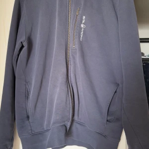 Mörkblå jacka från Sail Racing - Säljer en mörkblå jacka från Sail Racing med dragkedja framtill. Jackan har en enkel och stilren design med två fickor på sidorna och en liten logotyp på bröstet. Perfekt för en avslappnad stil.