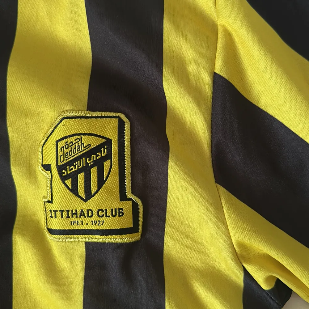 Säljer en gul och svart randig fotbollströja från Nike med korta ärmar. Tröjan har Ittihad Club-logga på bröstet och flera sponsorloggor. Perfekt för fotbollsfans som vill visa sitt stöd för laget.. T-paidat.