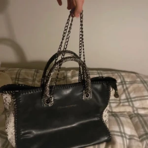 Svart handväska från Stella McCartney - Snygg svart handväska från Stella McCartney med kedjehandtag och ormskinnsmönstrade detaljer på sidorna. Perfekt för att ge din outfit en lyxig touch. ”Stella McCartney Python print faux hobo bag”  en väska som såldes i 2013 och är väldigt svår att få tag i nu då den inte säljs längre.