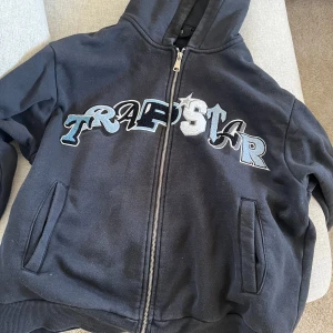 Svart hoodie från Trapstar - Säljer en svart hoodie från Trapstar med dragkedja och stort tryck på framsidan. Är storlek L och sitter bra på mig lite kort i midjan och jag är 186cm lång. Nyköpspris är 2300kr. Mitt pris kan diskuteras