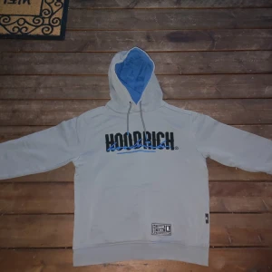 Grå hoodie från Hoodrich - Säljer en grå hoodie från Hoodrich med blått foder i huvan och tryck både fram och bak. Tröjan har långa ärmar och en klassisk känguruficka framtill. Perfekt för en avslappnad stil.