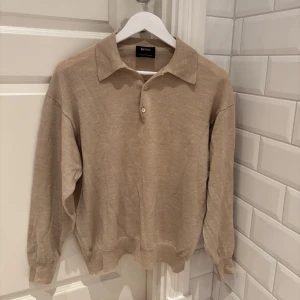 Beige merinoullströja från Hugo Boss - Säljer en stilren beige tröja från Hugo Boss i 100% merinoull. Tröjan har en klassisk krage med knappar och långa ärmar. Perfekt för en elegant och bekväm look. Storlek: Tror S, nypris: 1300kr