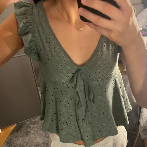 Grön blus med volangärmar - Säljer en söt grön blus med v-ringning och knytning framtill. Blusen har volangärmar och ett fint broderat mönster. Perfekt för en somrig look!