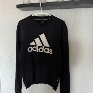 Svart sweatshirt från Adidas - Säljer en klassisk svart sweatshirt från Adidas med deras ikoniska logga i vitt på framsidan. Tröjan har långa ärmar och en rund halsringning. Perfekt för en avslappnad stil.