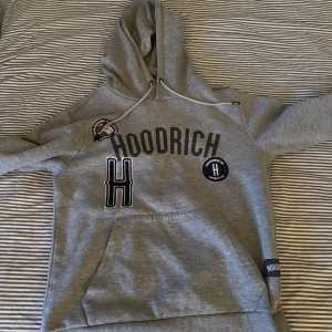 Grå hoodie från Hoodrich - Säljer en grå hoodie från Hoodrich med tryck på framsidan. Den har en stor ficka framtill och justerbar huva med dragsko. Perfekt för en avslappnad stil.